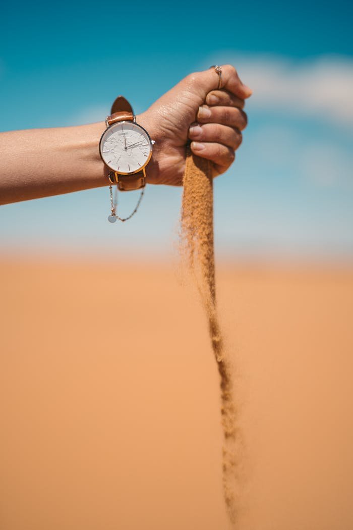 A watch on a hand pouring sand in the Moroccan desert under a clear blue sky.