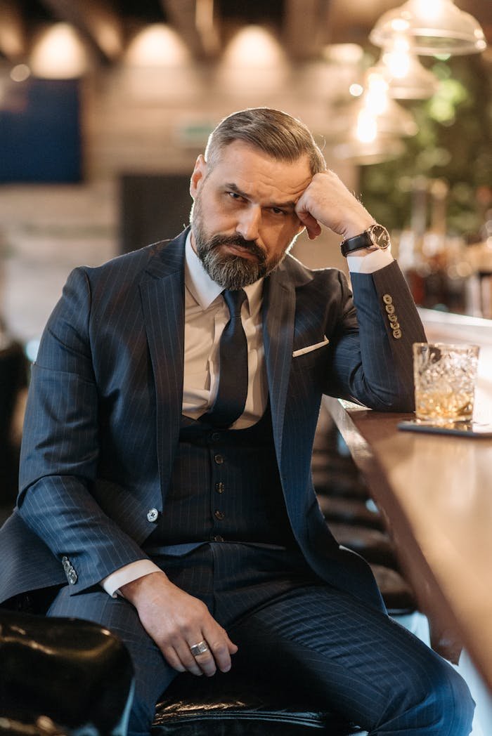 Sophisticated bearded man in blue suit sitting at bar counter, enjoying whiskey.